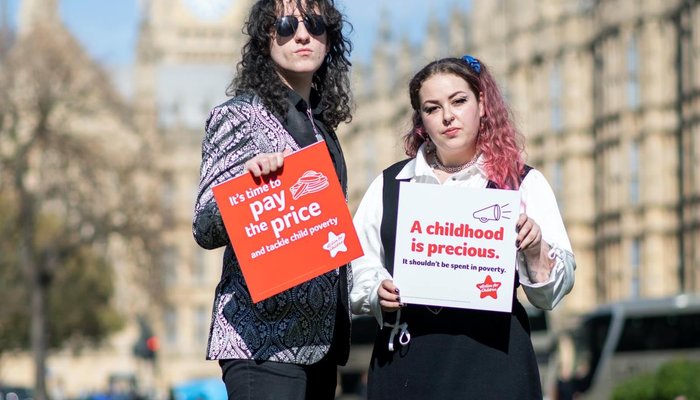 Two young campaigners outside of parliament holding Paying the Price placards with 'it's time to pay the price' and 'childhood is precious' messaging