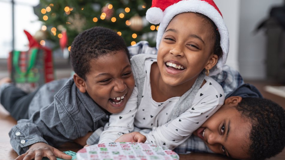 Three siblings laughing and laying on top of each other with Christmas gift presents in front of them a lit up tree in the background