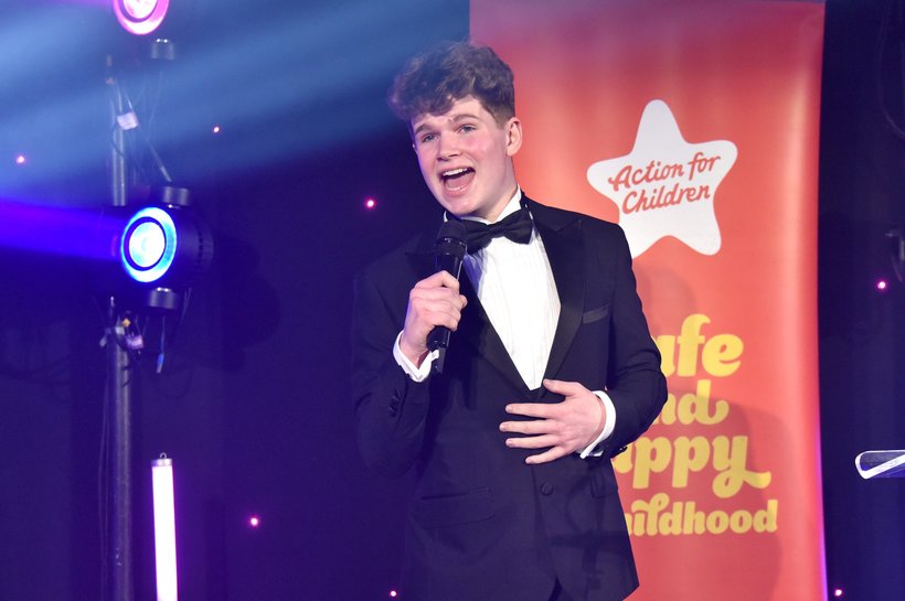 Young man wearing a suit and black bow tie on stage in front of an Action for Children pull up banner