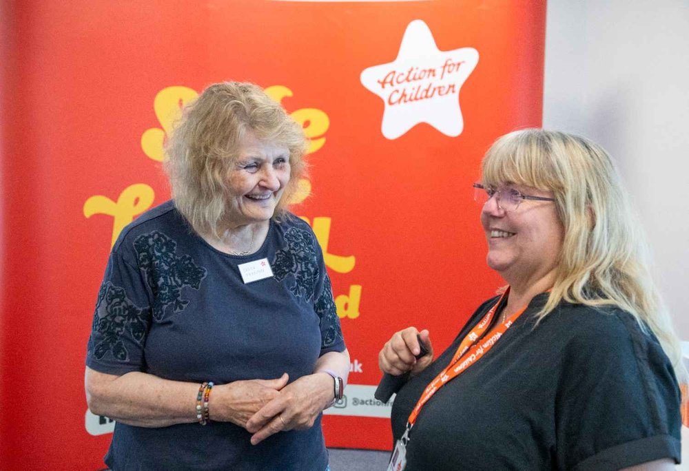 Two women chatting in front of an Action for Children backdrop