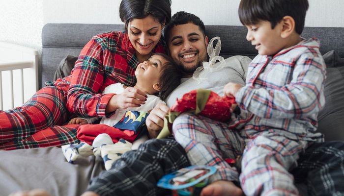 A family sitting on their bed during the Christmas period. The two young sons open presents as their parents watch smiling