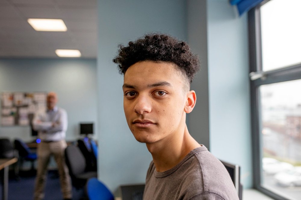 A teenage boy sits in class and looks at the camera as his tutor stands in the background. He is wearing casual clothing.
