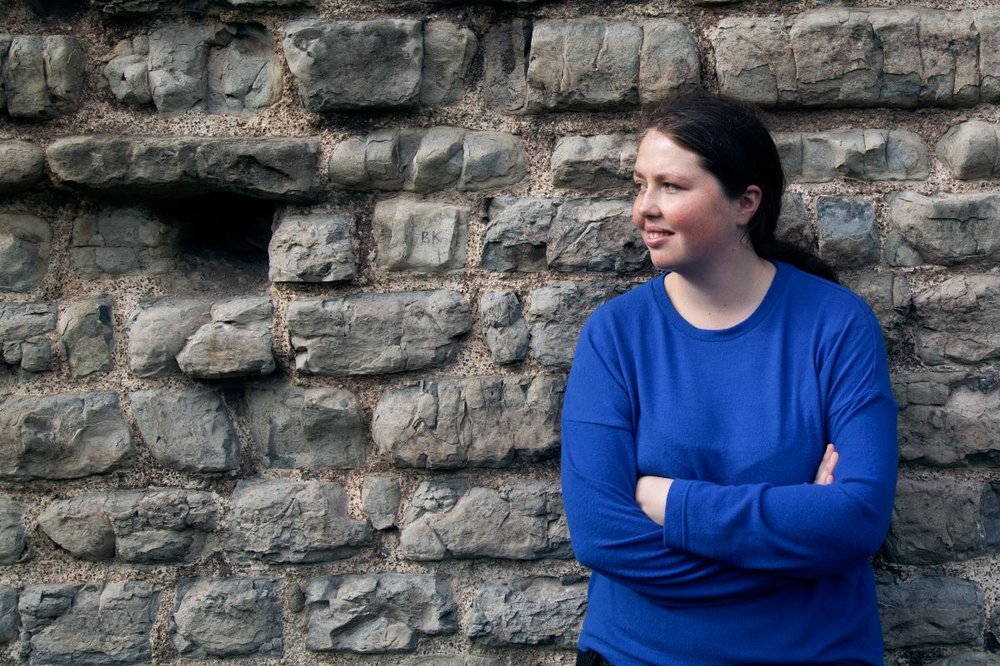 Angharad leaning against a wall smiling looking to the side