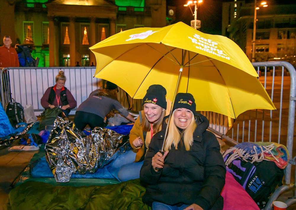 Two women at Boycott your Bed in their sleeping bags holding an umbrella - crop 4