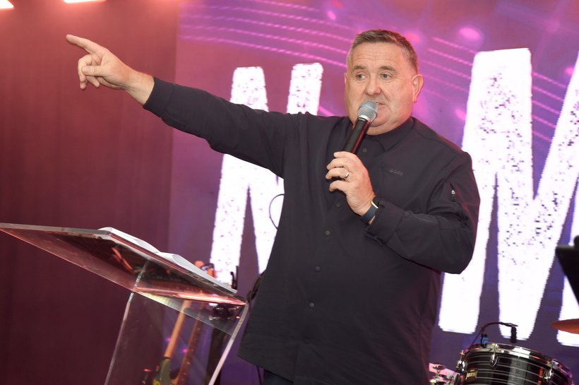 A man in a black shirt and jeans holding a microphone on stage pointing to the back of the room