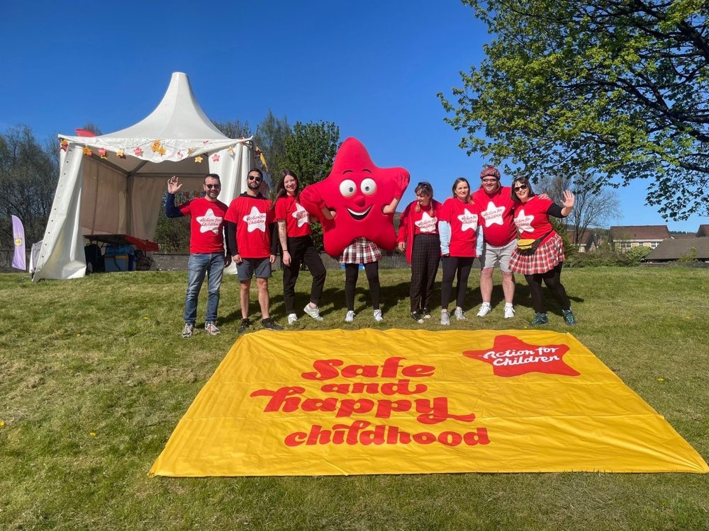 Group of people in Action for Children tshirts post with mascot Cosmo in front of a 'Safe and Happy Childhoods' sign