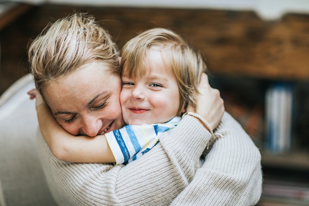 Happy mother hugging her small son at home, while boy is looking at camera
