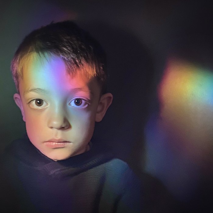 Head shot of a young boy with a neutral face. The light is refracting into a rainbow onto his face