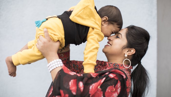 Jaspreet, wearing a black and red floral scarf, holds her baby son in a yellow and black tracksuit to her face. They smile and laugh looking at each other