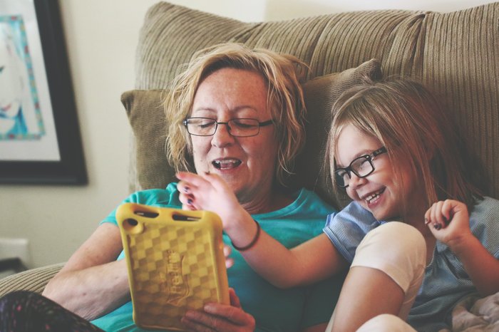 Grandmother and Granddaughter wearing glasses and sitting on the sofa together, looking at a phone and laughing