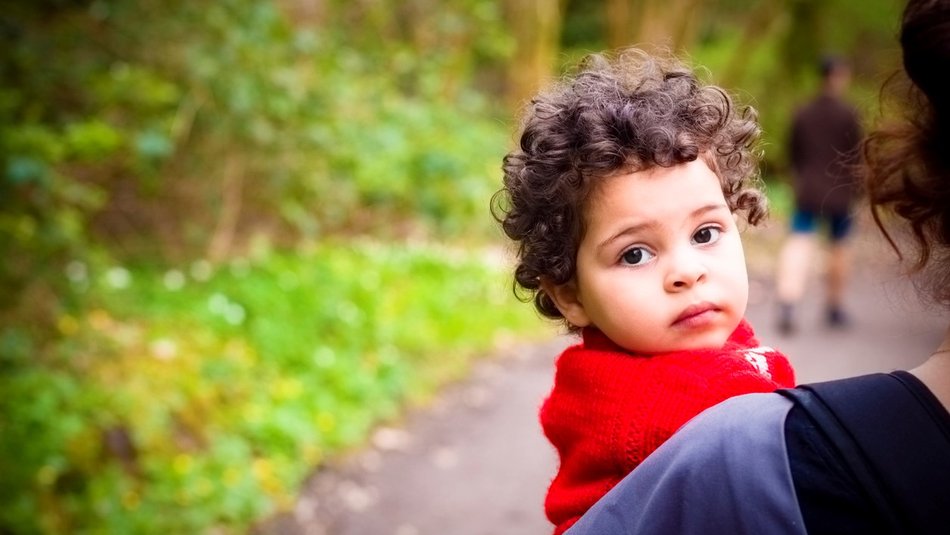Young girl in a red jumper being carried by her mother looking back at the camera