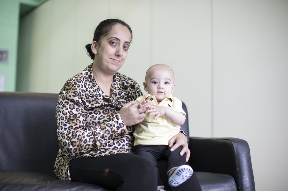 Shadia, a young woman in a anmal print shirt sits on a black sofa holding her baby son who also looking in to the camera