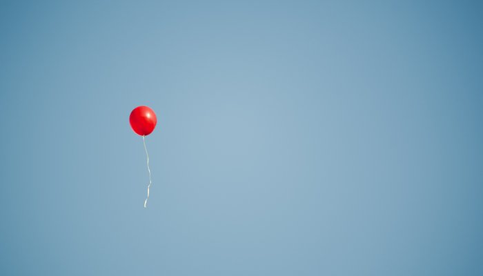 A red balloon with trailing string floats upwards against a clear blue sky