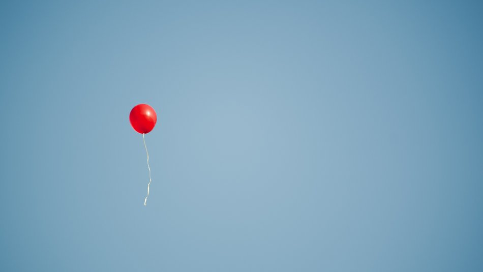 A red balloon with trailing string floats upwards against a clear blue sky