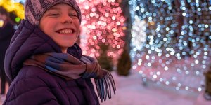 Teen boy outisde in warm clothes smiles at the camera. He's infront of a a background of glowing Christmas decorations