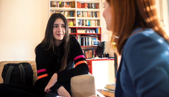 Teen girl sits talking opposite an adult woman who holds a clipboard