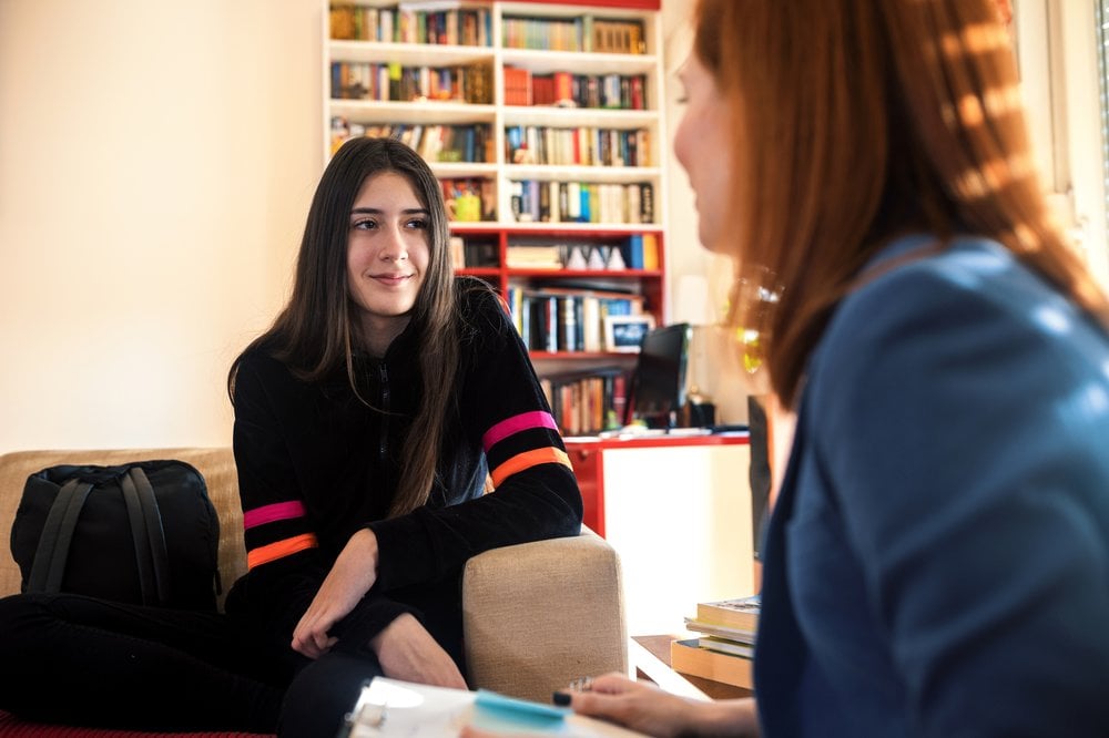 Teen girl sits talking opposite an adult woman who holds a clipboard