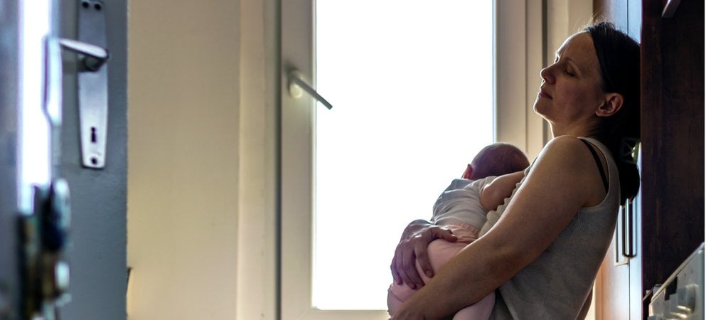 tired mother leans against kitchen wall, while rocking child to sleep