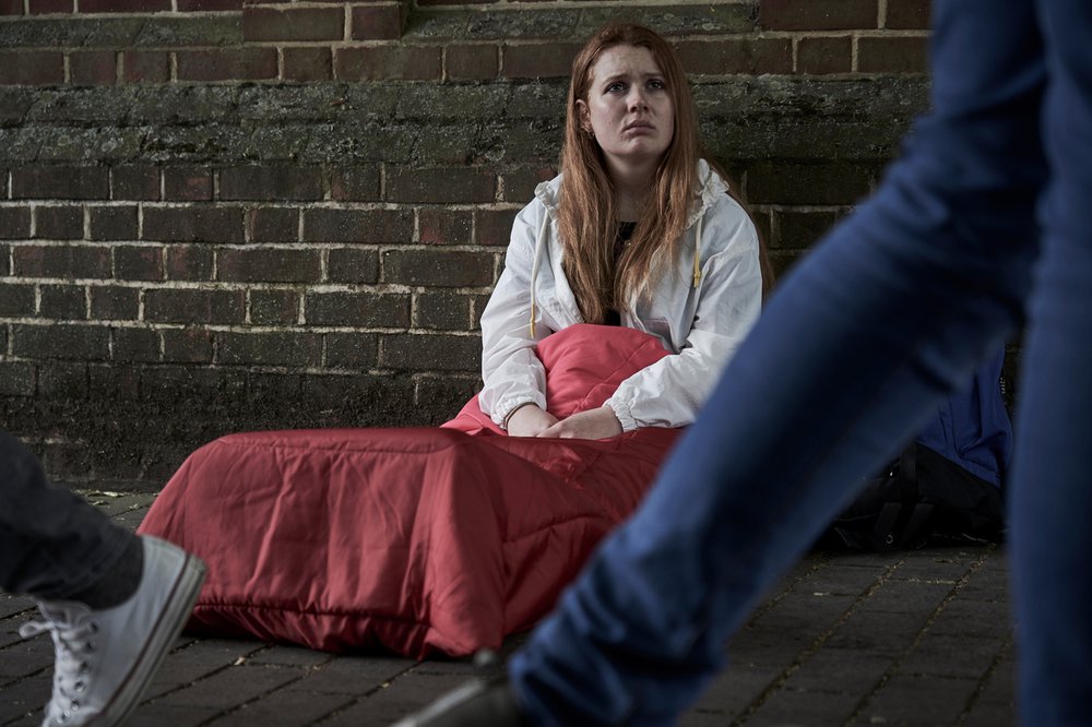 youg homeless girl sitting in sleeping back on street floor as people walk past.jpg