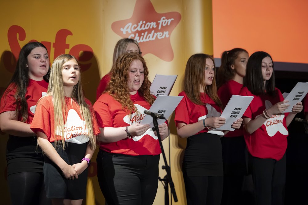 Young Carers Aloud Choir members, wearing Action for Children t-shirts, stand onstage singing at the Stephensons Award