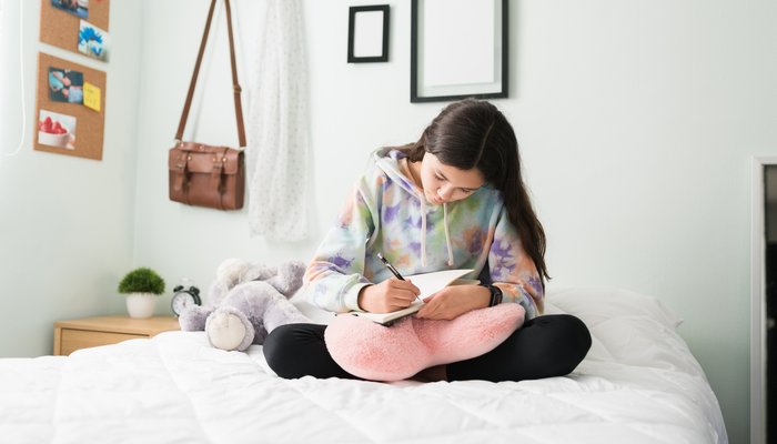 Young teen girl in her bedroom writing in a diary while sitting on her bed
