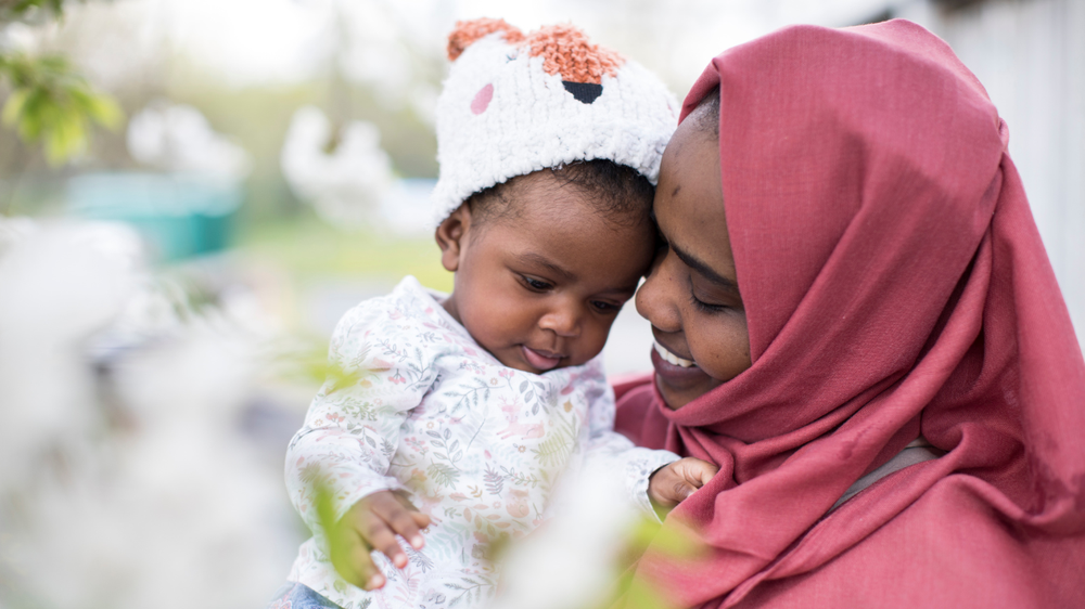 Samira, a woman in a red hijab smiles while looking at her baby daughter