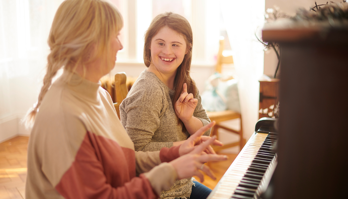 A woman and young girl sit at a wooden piano in a sunlight room