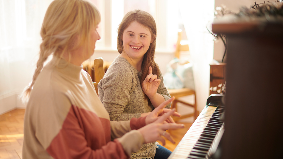 A woman and young girl sit at a wooden piano in a sunlight room