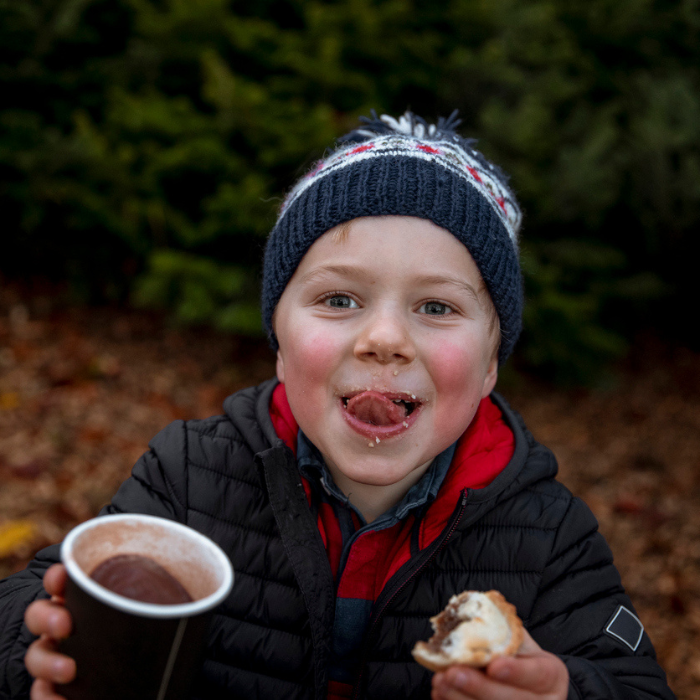 A young boy wearing warm clothing holding a warm drink and eating a mince pie